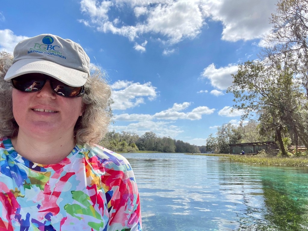 Photo of the Architect with hat, sunglasses, and SPF shirt in the foreground, beautiful blue-green river water with trees and party-cloudy blue skies in background.