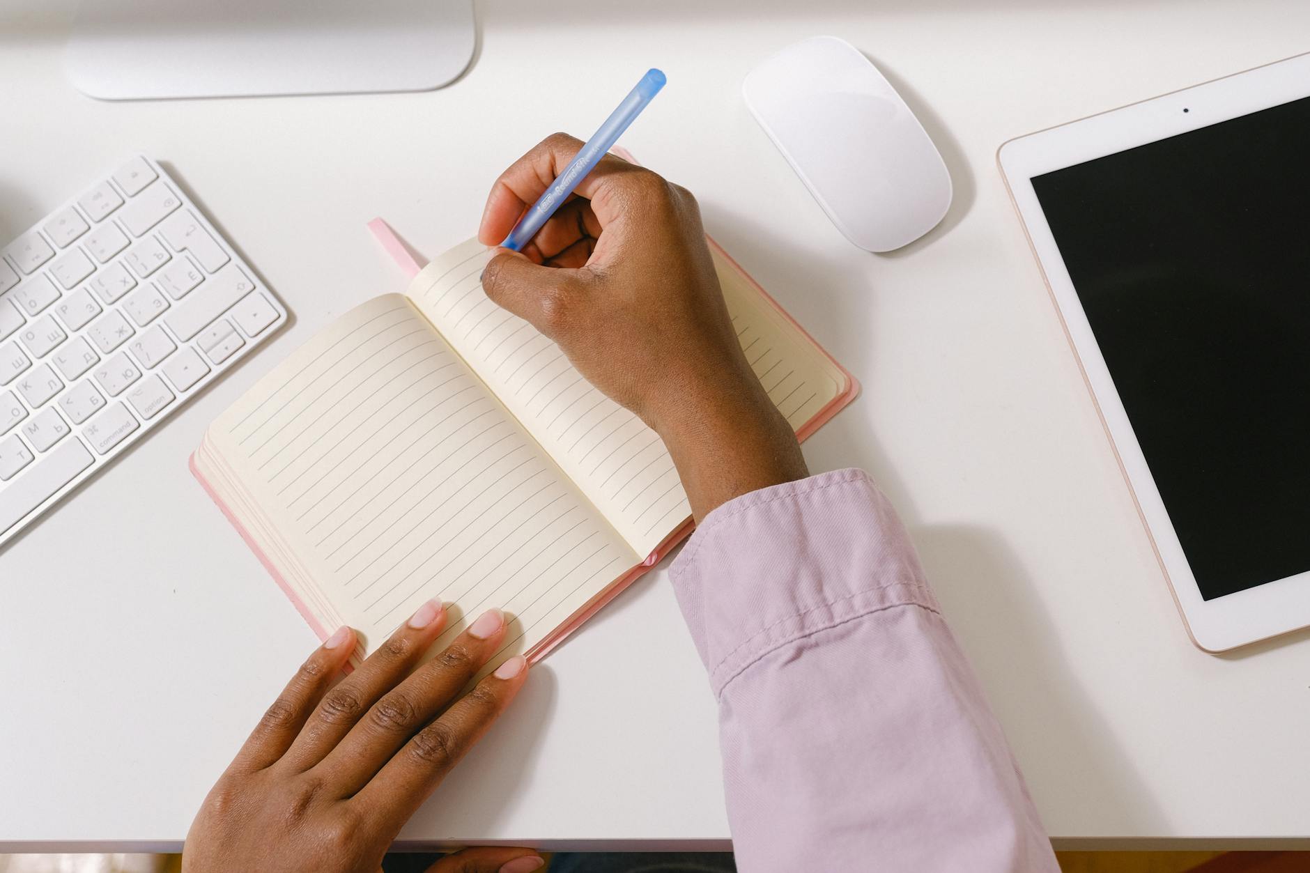 Photo of hands writing in a notebook on a white desk with a keyboard, mouse, and tablet nearby.