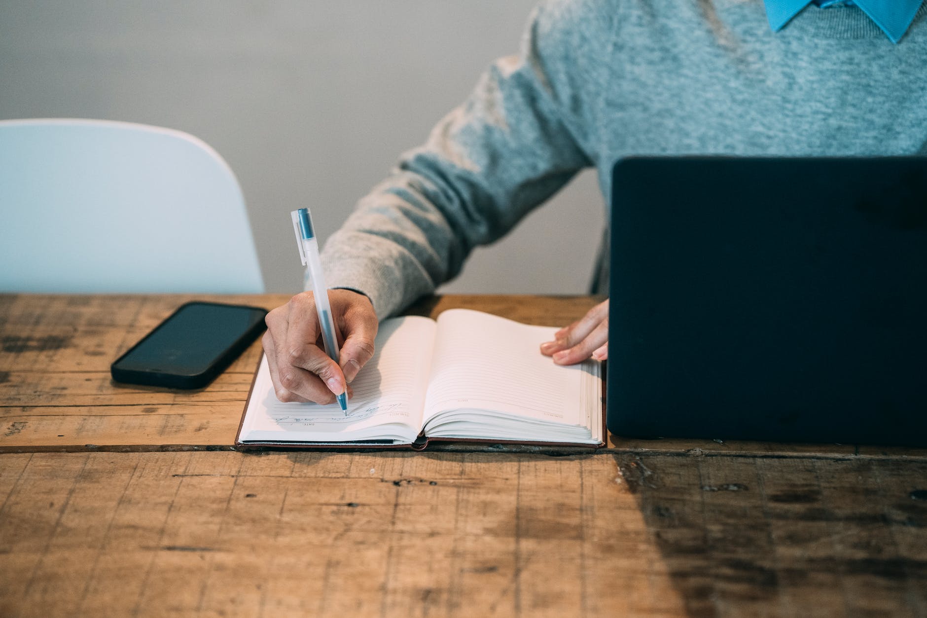 Photo of person writing in a notebook between a laptop computer and mobile phone on a wooden desk.