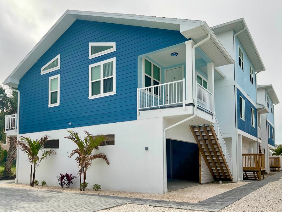 blue-and-white themed building with gable and parking below, front porch with long staircase to ground and taller duplex beyond